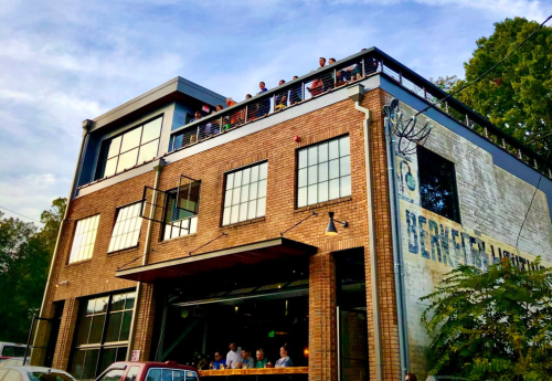 A brick building with large windows and a rooftop deck, where people are gathered, enjoying the outdoor space.