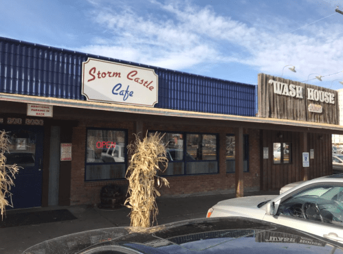 Exterior of Storm Castle Cafe with an "Open" sign, next to a rustic building labeled "Wash House." Clear blue sky above.