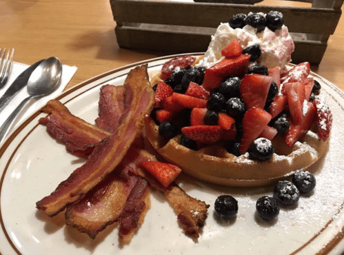 A plate featuring a waffle topped with strawberries, blueberries, whipped cream, and crispy bacon strips on the side.