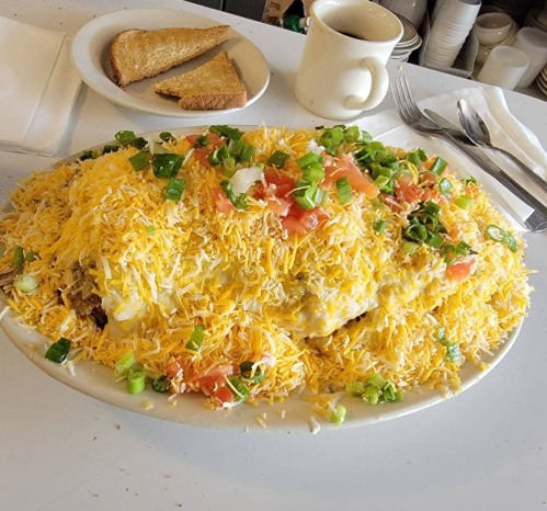 A large plate of cheesy nachos topped with tomatoes and green onions, served with toast and a cup of coffee in the background.