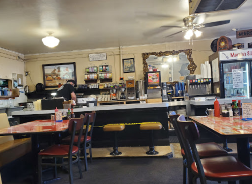 A cozy diner interior with red and black chairs, a counter, and a staff member preparing food in the background.