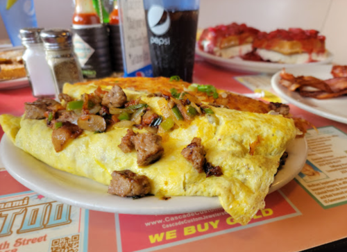 A hearty omelet filled with meat and green onions, served on a plate with sides of bacon and toast in the background.