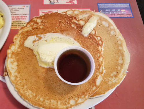 Two large pancakes with a pat of butter and a small cup of syrup on a red table.