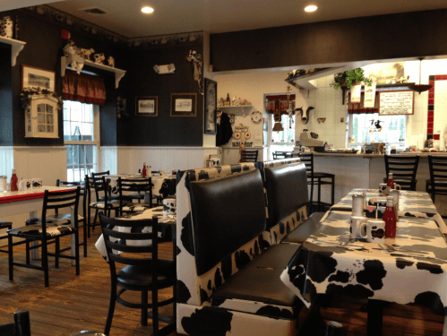 Cozy diner interior with cow-print decor, black chairs, and tables set for dining. A kitchen area is visible in the background.