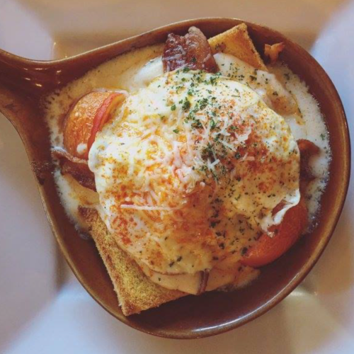 A baked dish with a poached egg, toast, tomatoes, and cheese, garnished with herbs in a brown bowl.