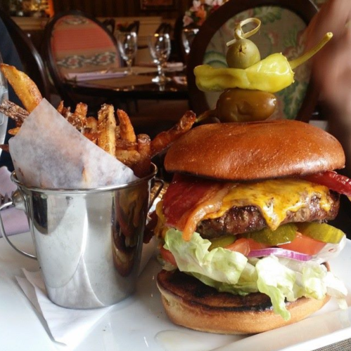 A juicy cheeseburger with lettuce, tomato, and pickles, served with a side of crispy fries in a metal cup.