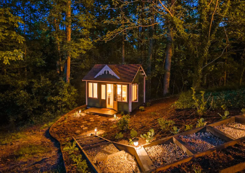 A cozy cabin nestled in the woods, illuminated by soft lights at dusk, surrounded by trees and a gravel path.