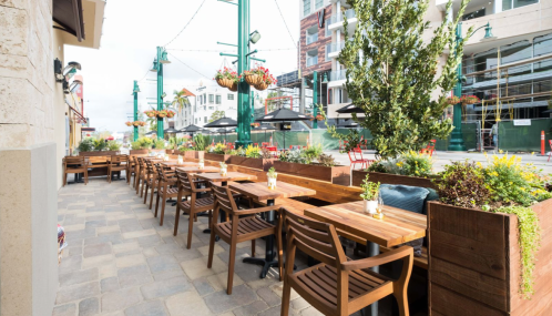 Outdoor dining area with wooden tables, chairs, and planters, set against a backdrop of buildings and construction.