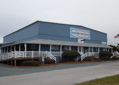 Blue historical building with a sign reading "Assembly Building" and an American flag, surrounded by greenery.