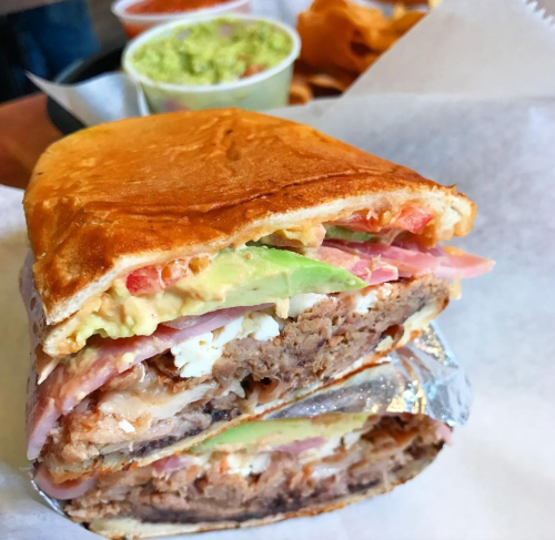 A stacked sandwich with layers of meat, avocado, tomato, and cheese, served with sides of guacamole and salsa.