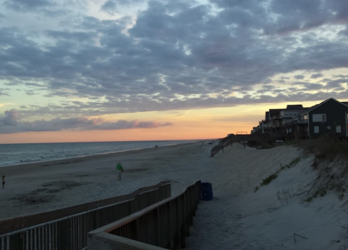 A serene beach at sunset, with clouds in the sky and houses lining the shore. Gentle waves lap at the sand.