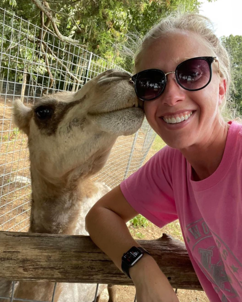 A woman in sunglasses smiles as a camel leans in for a kiss at a petting zoo.