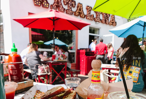 Outdoor dining scene with colorful umbrellas, people enjoying food, and a bottle of hot sauce in the foreground.