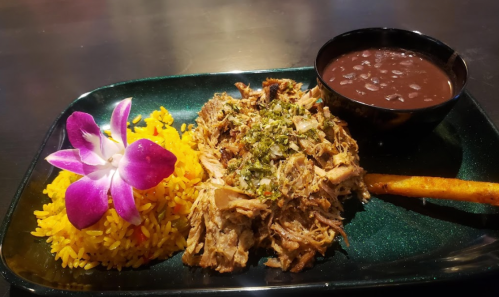A plate featuring shredded meat topped with herbs, yellow rice, black beans, a plantain, and a purple orchid garnish.