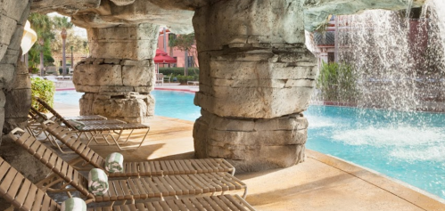 A serene pool area with stone pillars, lounge chairs, and a waterfall, surrounded by greenery and a clear blue sky.