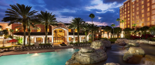 A luxurious hotel with palm trees, a pool, and a waterfall, set against a twilight sky.