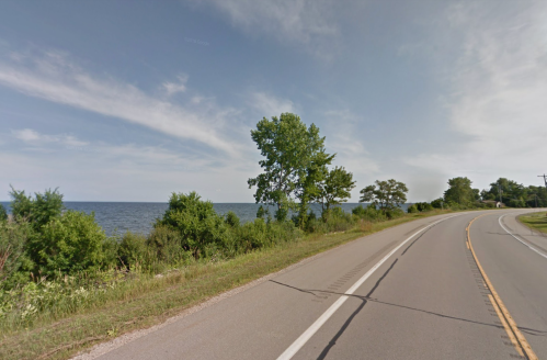 A scenic view of a road alongside a lake, with trees and greenery lining the shore under a blue sky.