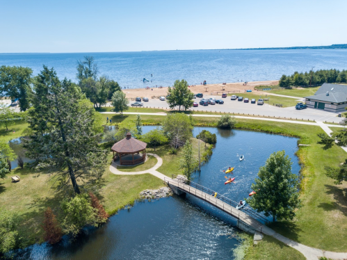 Aerial view of a beach park with a gazebo, pond, and kayakers, overlooking a calm lake under a clear blue sky.