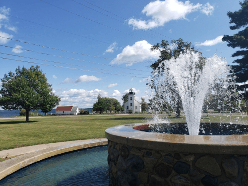 A fountain sprays water in a park with a lighthouse and trees in the background under a blue sky with clouds.