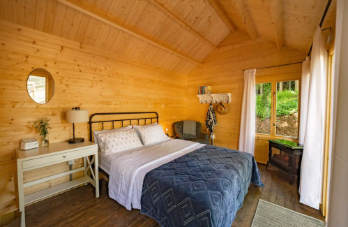 Cozy wooden bedroom with a bed, nightstand, and large window, featuring natural light and rustic decor.