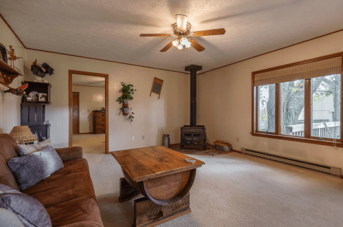 Cozy living room with a brown sofa, wooden coffee table, ceiling fan, and a wood stove near a large window.