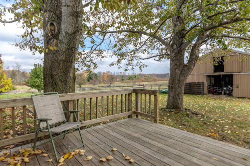 A wooden deck with a chair, surrounded by trees and autumn leaves, overlooking a barn and open fields.