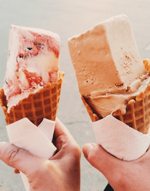 Two hands holding ice cream cones: one with strawberry swirl and the other with caramel flavor, both in waffle cones.