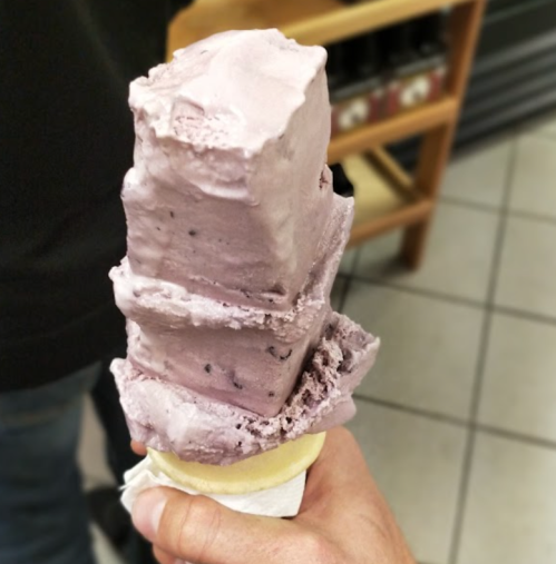 A hand holding a tall ice cream cone with three stacked scoops of purple ice cream.