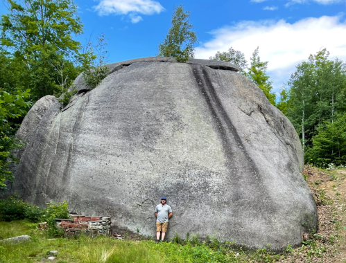 A person stands beside a large, smooth rock formation surrounded by greenery and blue sky.