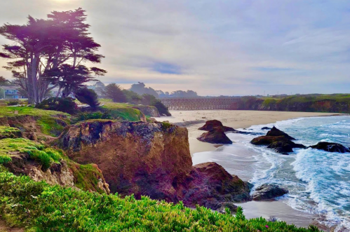 Coastal view featuring rocky cliffs, sandy beach, and a wooden pier under a cloudy sky. Lush greenery in the foreground.