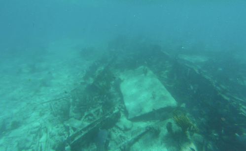 Underwater scene showing a sunken shipwreck surrounded by sand and marine life in a blue ocean environment.