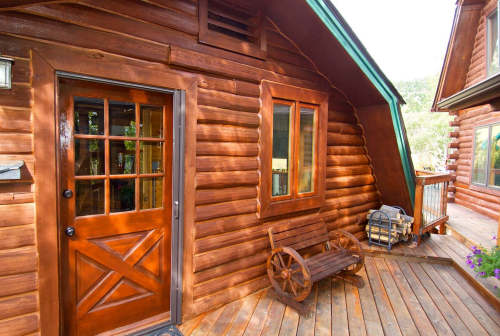 A wooden log cabin entrance with a door, window, and a bench on a deck surrounded by greenery.