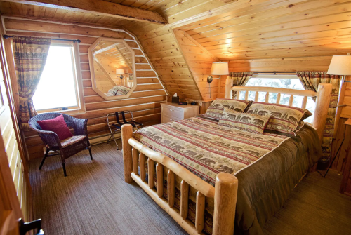 Cozy wooden bedroom with a log bed, patterned bedding, and large windows, featuring rustic decor and warm lighting.