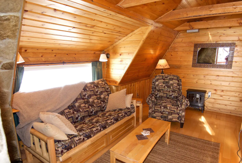 Cozy wooden cabin interior with a sofa, coffee table, and warm lighting. Rustic decor and a sloped ceiling create a welcoming atmosphere.