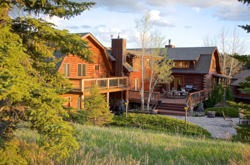 A rustic wooden cabin surrounded by trees, featuring a spacious deck and stone patio in a scenic outdoor setting.