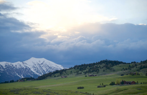 A scenic landscape featuring snow-capped mountains, rolling green hills, and a cloudy sky at dawn.