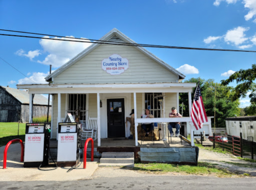 A small country store with an American flag, featuring two people sitting outside and vintage gas pumps in front.