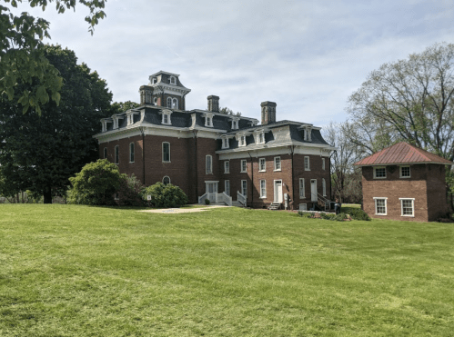 A large, historic brick mansion with a tower, surrounded by green grass and trees, with a smaller building nearby.