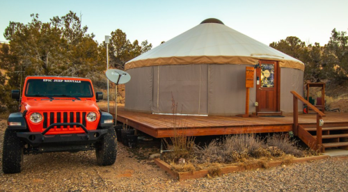 A red Jeep parked next to a wooden deck yurt surrounded by trees in a rural setting.