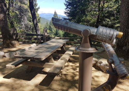 A telescope overlooks a scenic view, with a wooden picnic table nearby, surrounded by trees and mountains in the background.