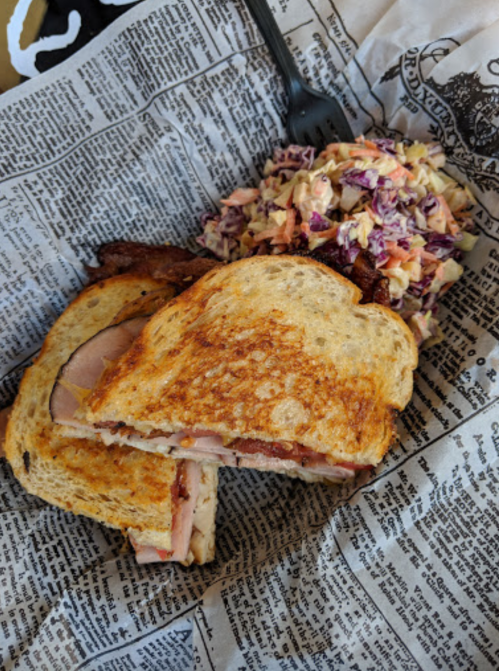 A toasted sandwich with layers of meat, served with a side of colorful coleslaw on newspaper print.