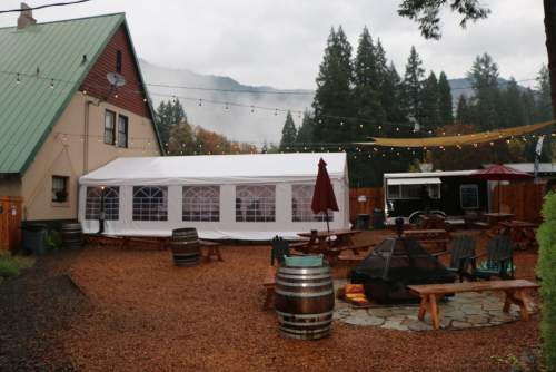 A cozy outdoor space with a white tent, wooden tables, and string lights, surrounded by trees and mountains in the background.