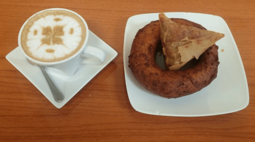 A cup of cappuccino beside a plate with a donut and a triangular pastry on a wooden table.