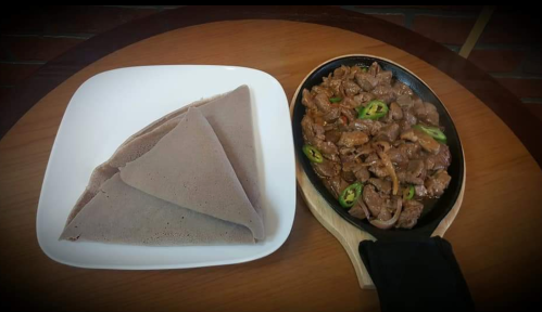 A plate of injera next to a sizzling dish of spicy beef with green peppers on a wooden board.