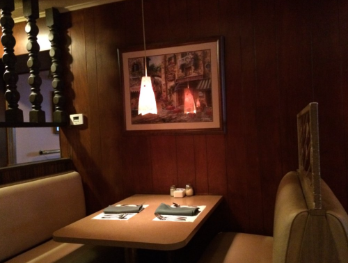 Cozy restaurant booth with wooden walls, a framed painting, and a pendant light hanging above a table set for two.