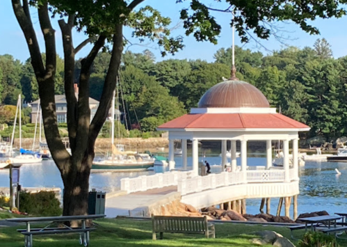 A gazebo by the water with boats in the background, surrounded by trees and a clear blue sky.