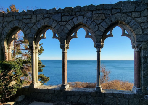 A stone archway with three pointed arches frames a serene view of the ocean and clear blue sky.