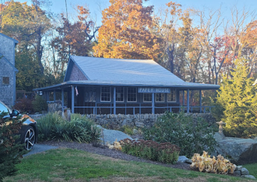 A quaint wooden house with a sign reading "Paper House," surrounded by trees and a stone wall.