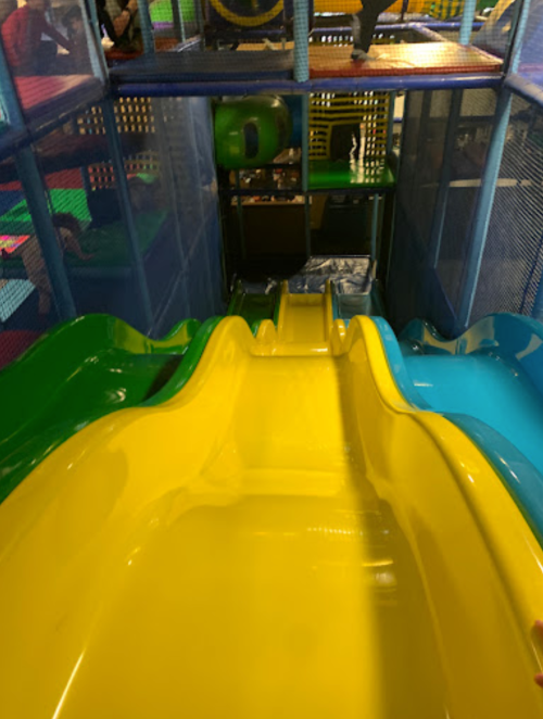 Colorful indoor play structure featuring yellow and blue slides, with a netted area and soft play surfaces visible.