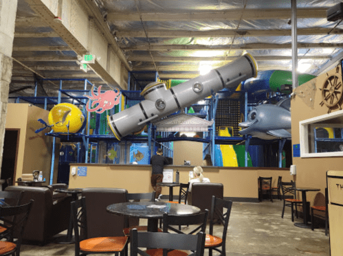 Indoor play area with colorful slides and structures, visible from a dining area with tables and chairs.